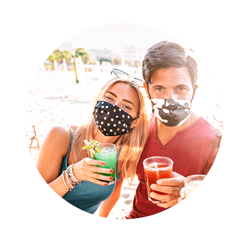 A man and a woman on a sunny exotic beach holding cocktails whilst wearing face masks
