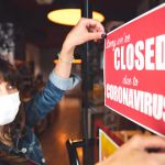 A woman with dark hair and wearing a face mask puts up a sign on a shop door that reads "Sorry we are closed due to coronavirus"