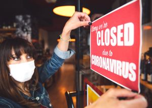 A woman with dark hair and wearing a face mask puts up a sign on a shop door that reads "Sorry we are closed due to coronavirus"