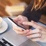 Close up of a woman using her mobile phone whilst sat at a laptop with a coffee