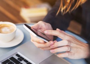 Close up of a woman using her mobile phone whilst sat at a laptop with a coffee