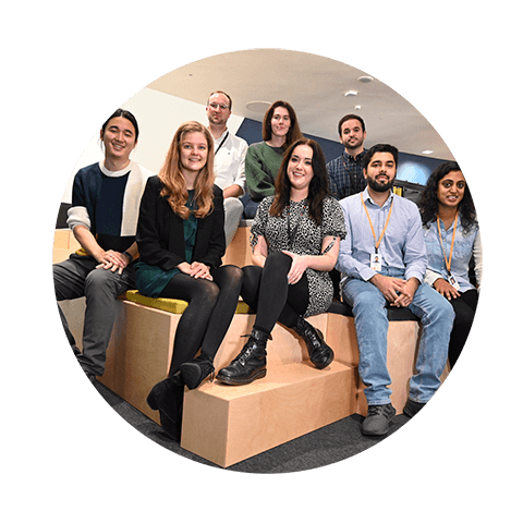 A smiling team of young men and women wearing lanyards sat in a modern corporate office space